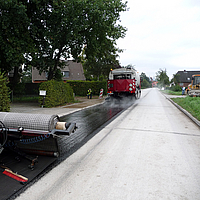 Une machine à terme se déplace en avant, suivie par l'application de l'armature d'asphalte SamiGrid® sur la route.