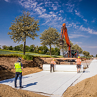 Une pelleteuse équipée d'une traverse de pose déroule le géotextile Stabilenka sur le chantier